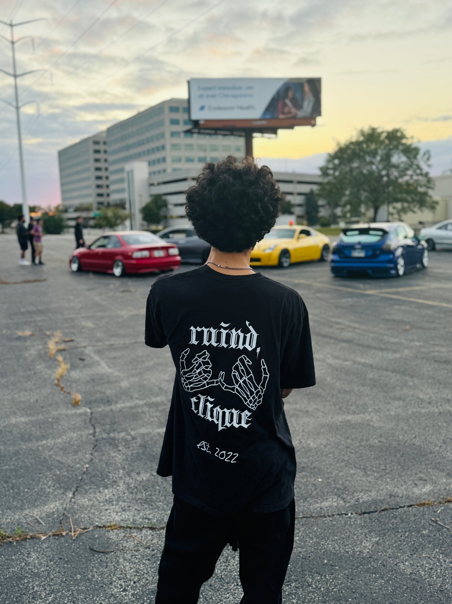 Black t-shirt from the back, featuring the 'Ruind Clique' club name and two skeleton hands linked together. The scene is set in front of a lineup of stylish cars, adding a bold and urban aesthetic.