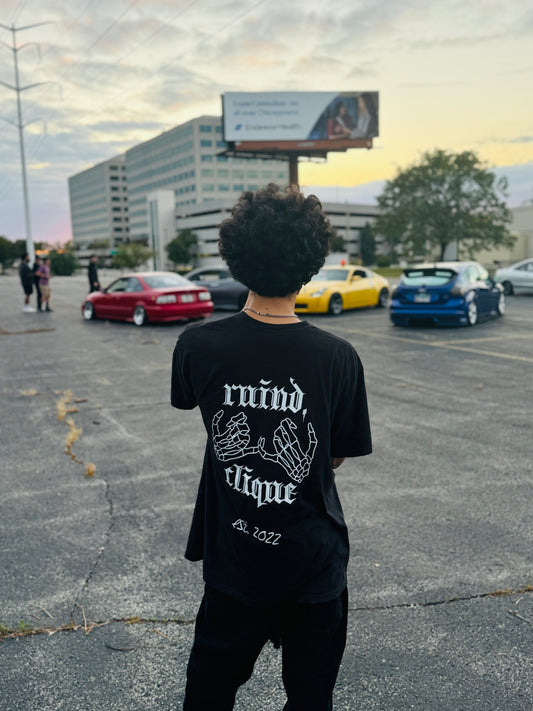 Black t-shirt from the back, featuring the 'Ruind Clique' club name and two skeleton hands linked together. The scene is set in front of a lineup of stylish cars, adding a bold and urban aesthetic.